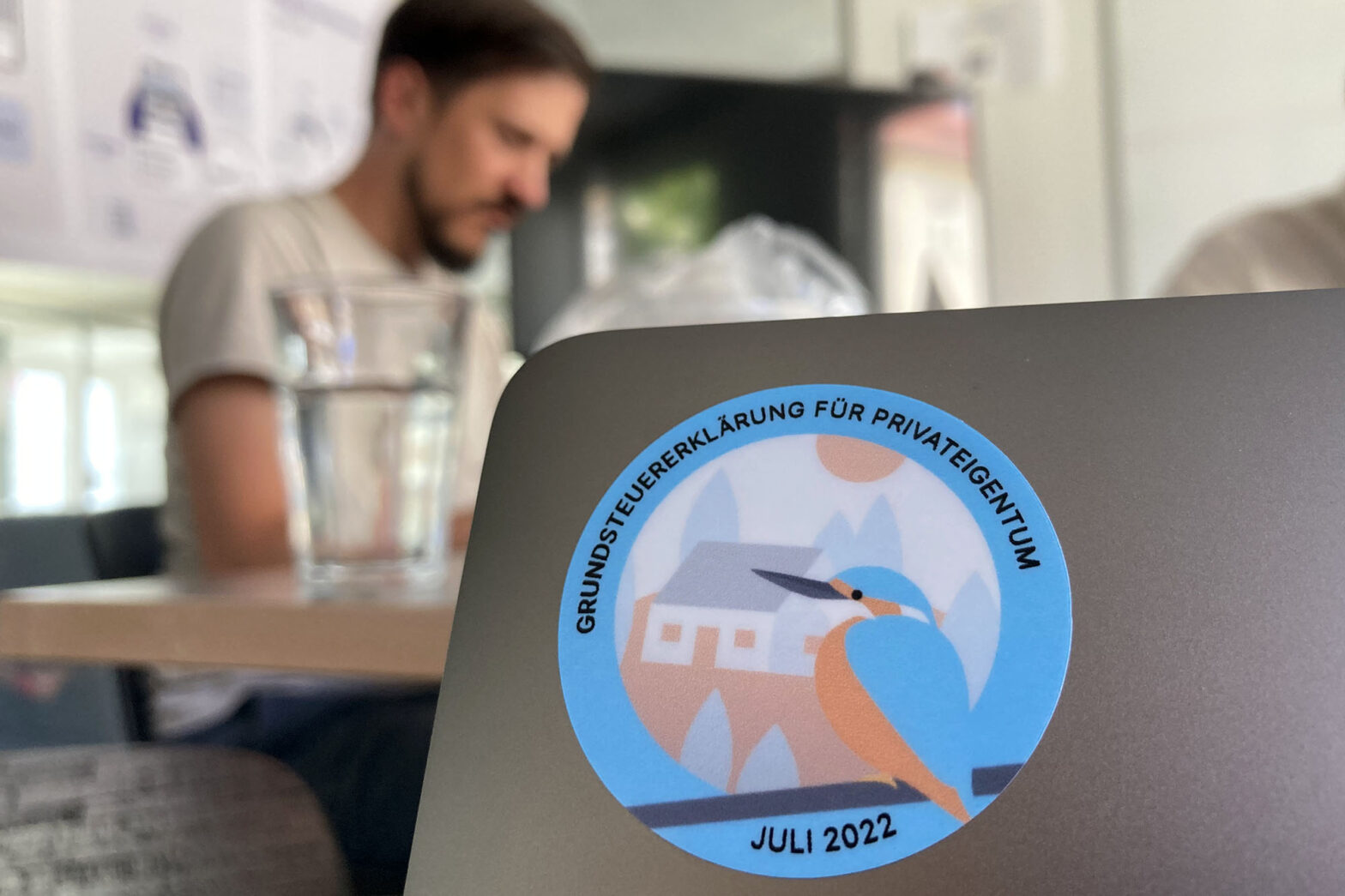 A circular sticker on a laptop computer with a common kingfisher in front of a house that says: private property tax declaration – July 2022 in German and a young man sitting behind the computer