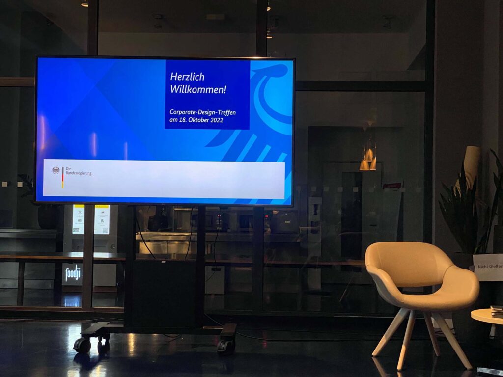 A dark space with a small lounge chair and table standing on the right and a big screen on the left; it shows a slide saying ‘Welcome! Corporate Design Meeting on 18 October 2022 – the Federal government‘