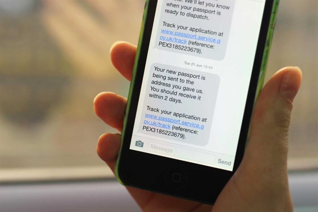 A person’s hand holding a smartphone showing two text messages, the most recent one saying: ‘Your new passport is being sent to the address you gave us. You should receive it within 2 days. Track you application at www.passport.service.gov.uk/track’