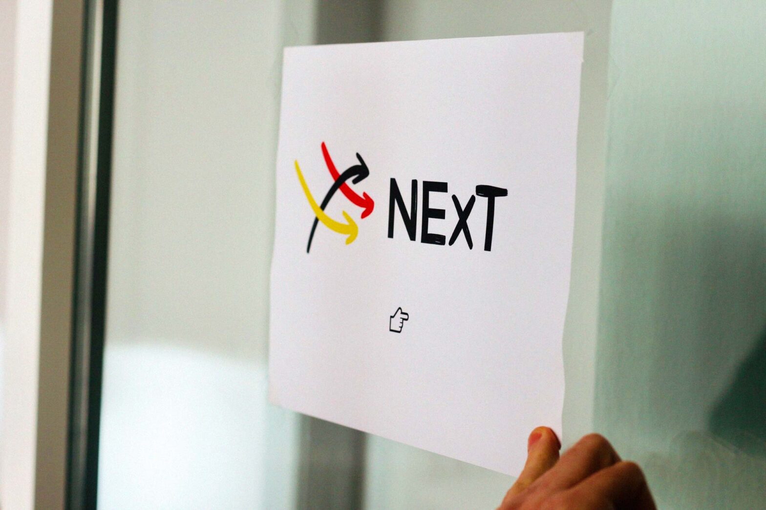 A sign with the logo of NExT with 3 hand-drawn arrows in black, red and yellow taped to a glass door with a hand touching it