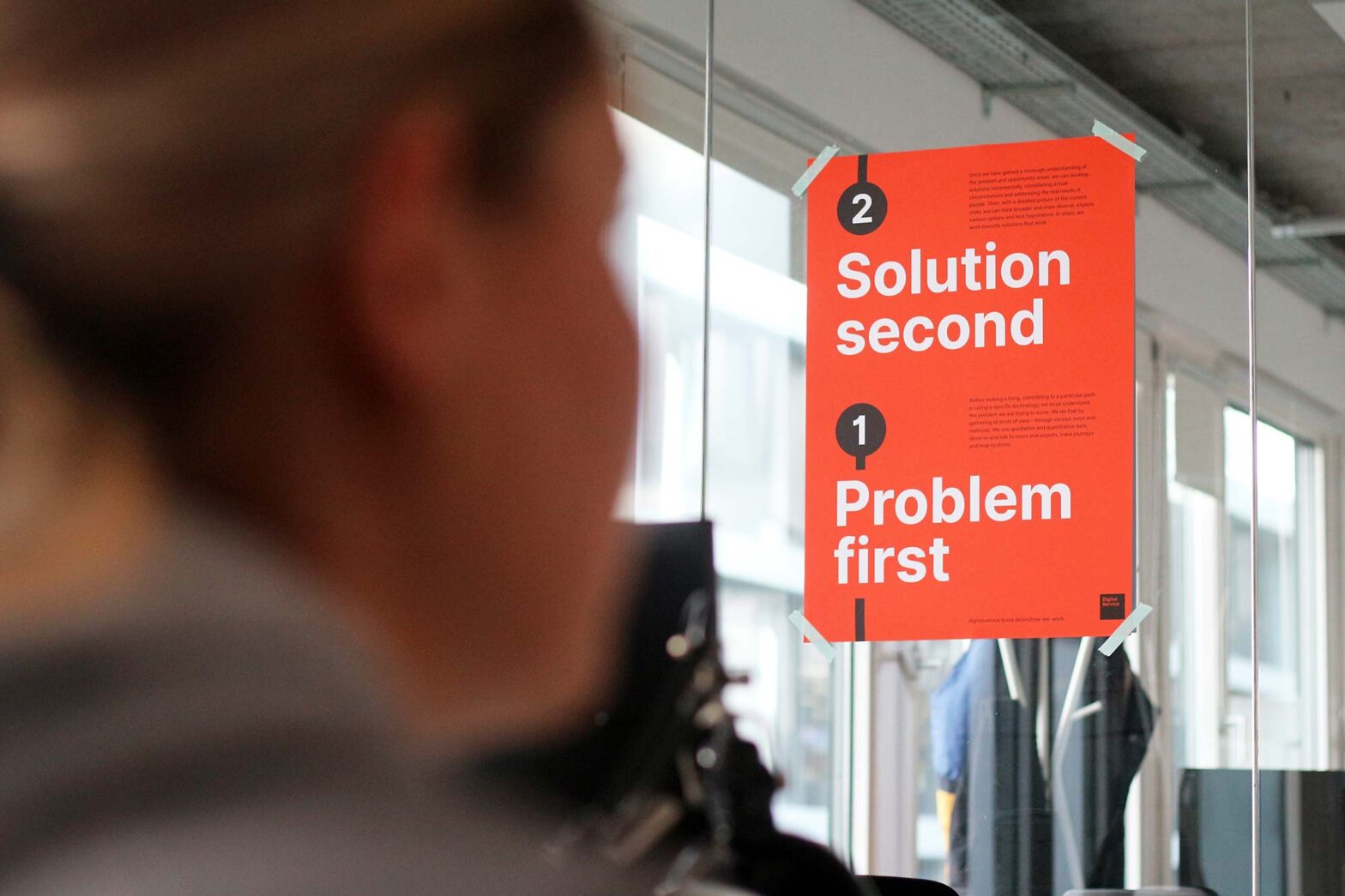 A bright orange poster stuck to a glass wall in a modern office space with a person sitting somewhat in front of it. The poster shows 2 steps in the style of underground map with numbers: 2 solution second 1 problem first further additional text is placed next to the numbers.