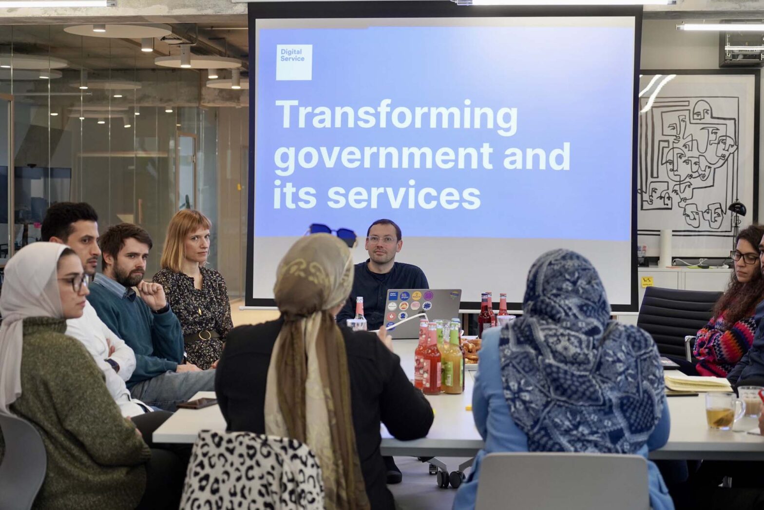A group of people of different skin tones sitting around a larger table in a modern office space with glass and concrete, a projected slide behind them says: “Digital Service — Transforming government and its services”; some people wear office casual clothes, other contemporary Arabic clothes with headscarves