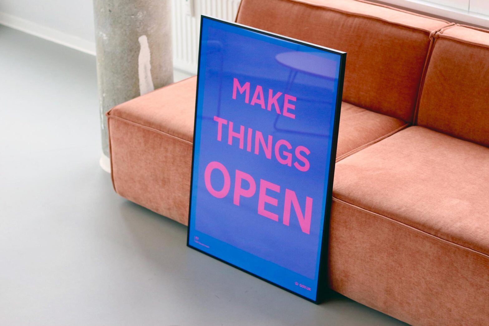 A bright blue poster with big pink letters in a slim black frame that reads ‘Make things open’, it leans on a brown sofa in a modern grey-white office space with concrete walls
