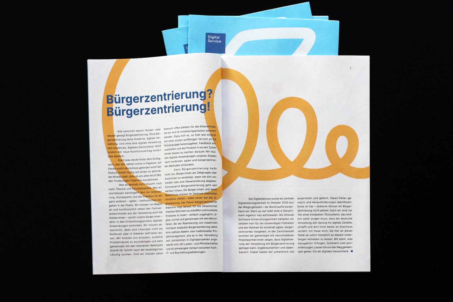 An open booklet with a lot of text, a headline and a curly line in the background. The headline says “Citizen-centricity? Citizen-centricity!” in German. Behind are further booklets piled up revealing a square logo that says “Digital Service”.