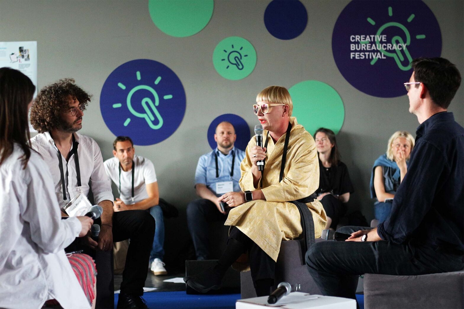 A group of 4 people with microphones and lanyards are sitting on stools, discussing – 4 more people who look at them sit behind them, leading at the wall with graphical applications of logos of ‘Creative Bureaucracy Festival’ – all people have lighter skin tones