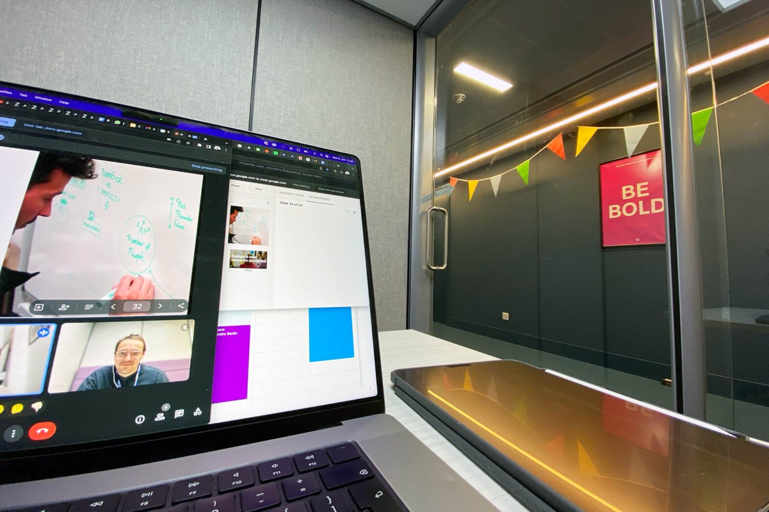 A closeup of a laptop showing multiple screens during a video call including a youngish white man with glasses sitting in a cubicle; outside of it hangs a pink poster saying ‘Be bold’ in big letters and some colourful bunting