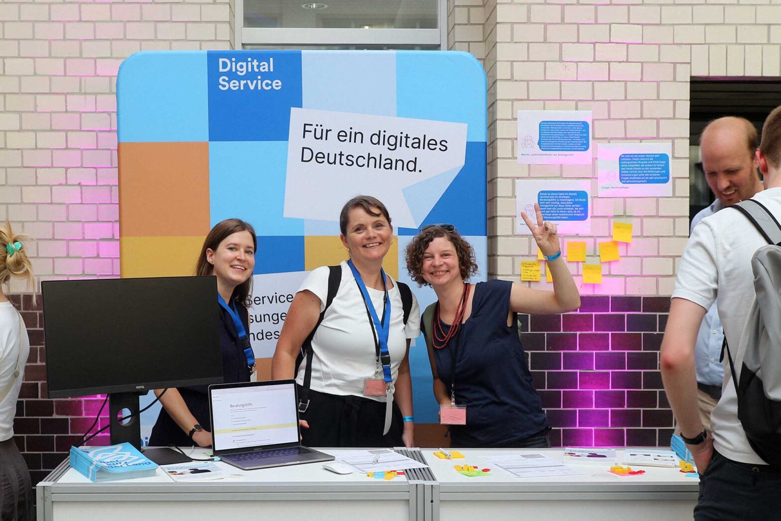 Three women with lighter skin standing at booth smiling – behind them is pop-up display with the logo of Digital Service and a big headline saying: “For a digital Germany”