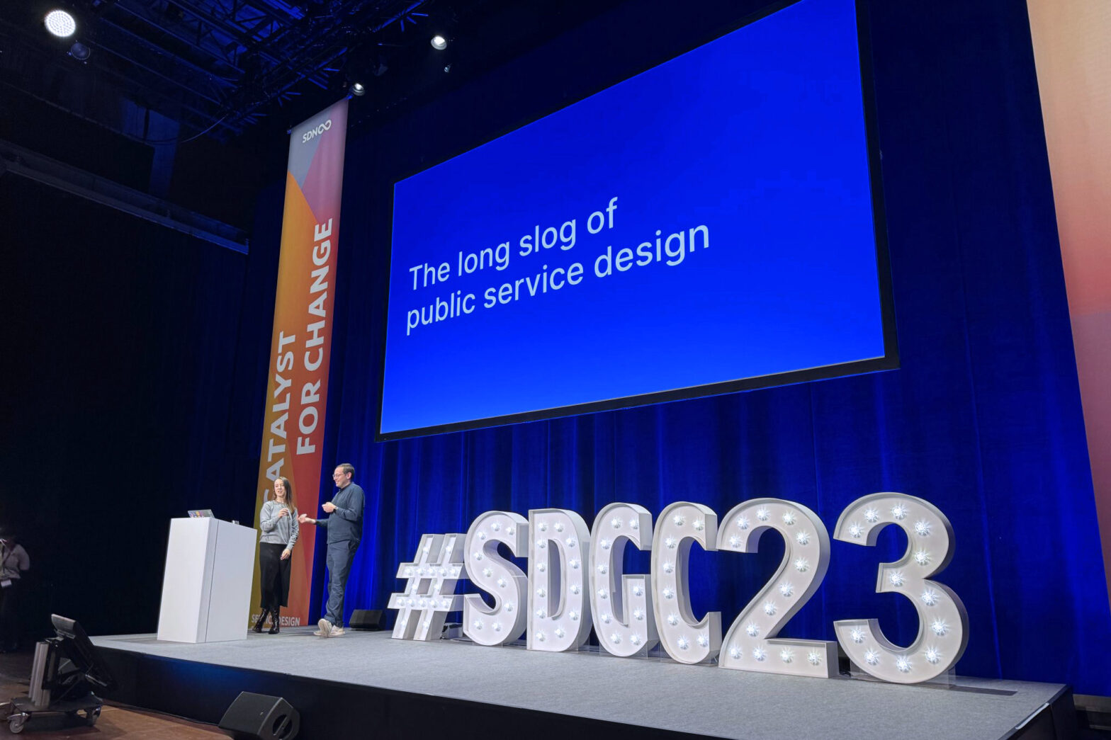 A white woman and a white man are on a big stage, behind them is a banner saying ‘catalyst for change’ and a projected slide saying ‘the long slog of public service design’, next to them are huge letters and numbers: SDGC23
