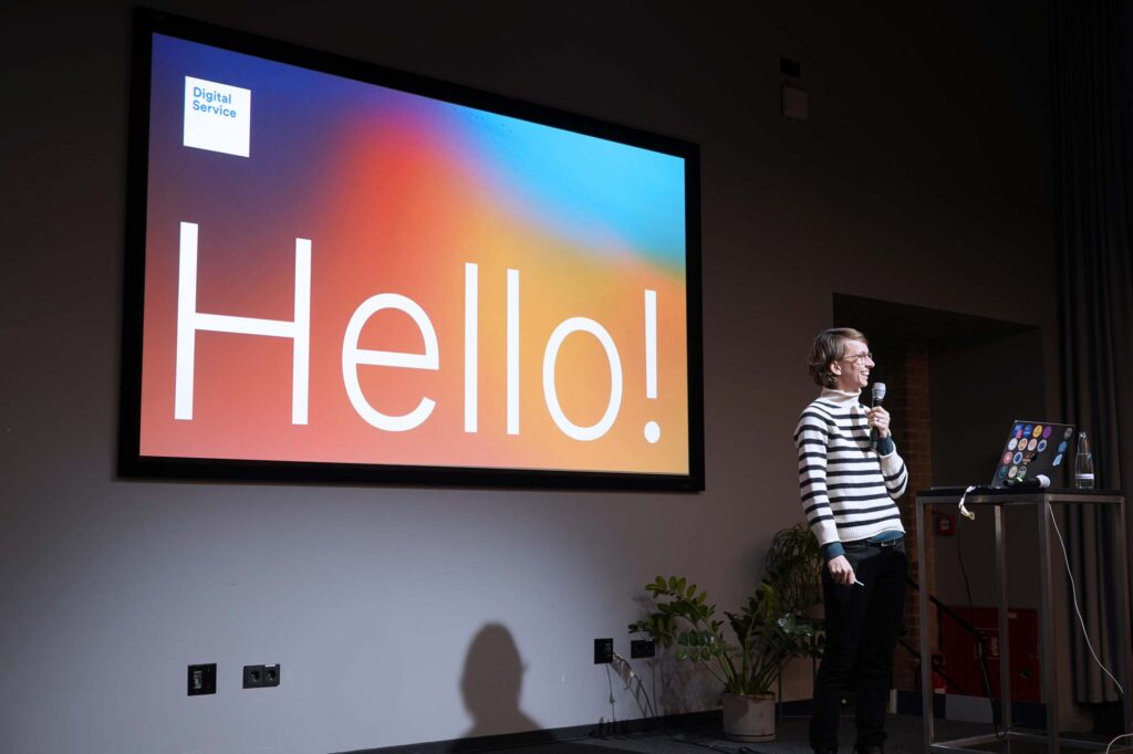 A woman with light skin and short hair in a striped wool jumper holding a microphone while standing on a stage with lecturn with a computer next to her, a slide is projected big behind her saying “Hello”