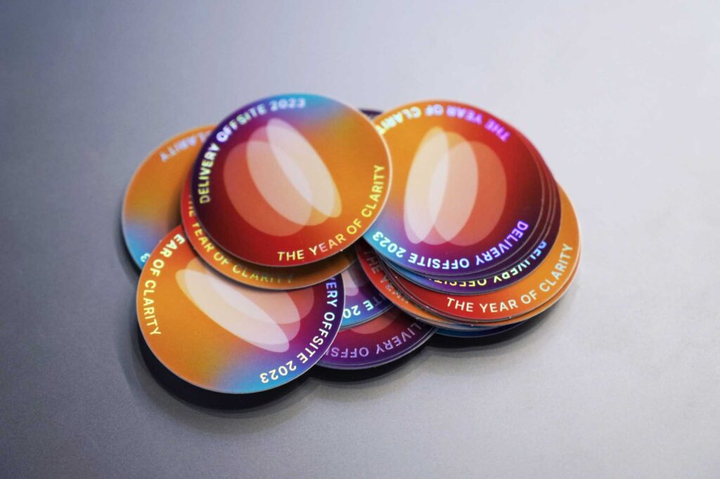 A pile of colourful round stickers with shiny reflective foil saying: “DELIVERY OFFSITE 2023 – THE YEAR OF CLARITY”