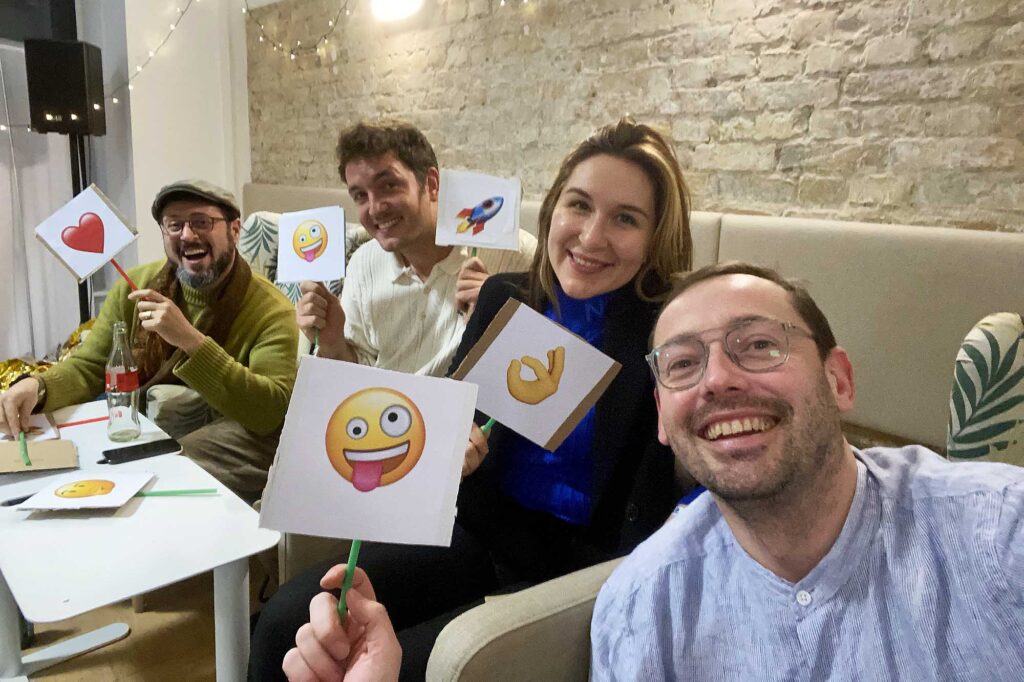 A selfie of group of 4 white people – 3 men and 1 woman, sitting on sofa, smiling into the camera, holding small signs showing emojis: a heart, a rocket, an ok hand, and a zany face 