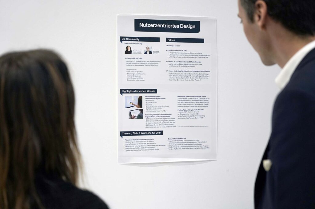 Two people in formal clothes standing in front of a poster with lots of text and a few small pictures – the poster’s headline says in German ‘user-centred design’