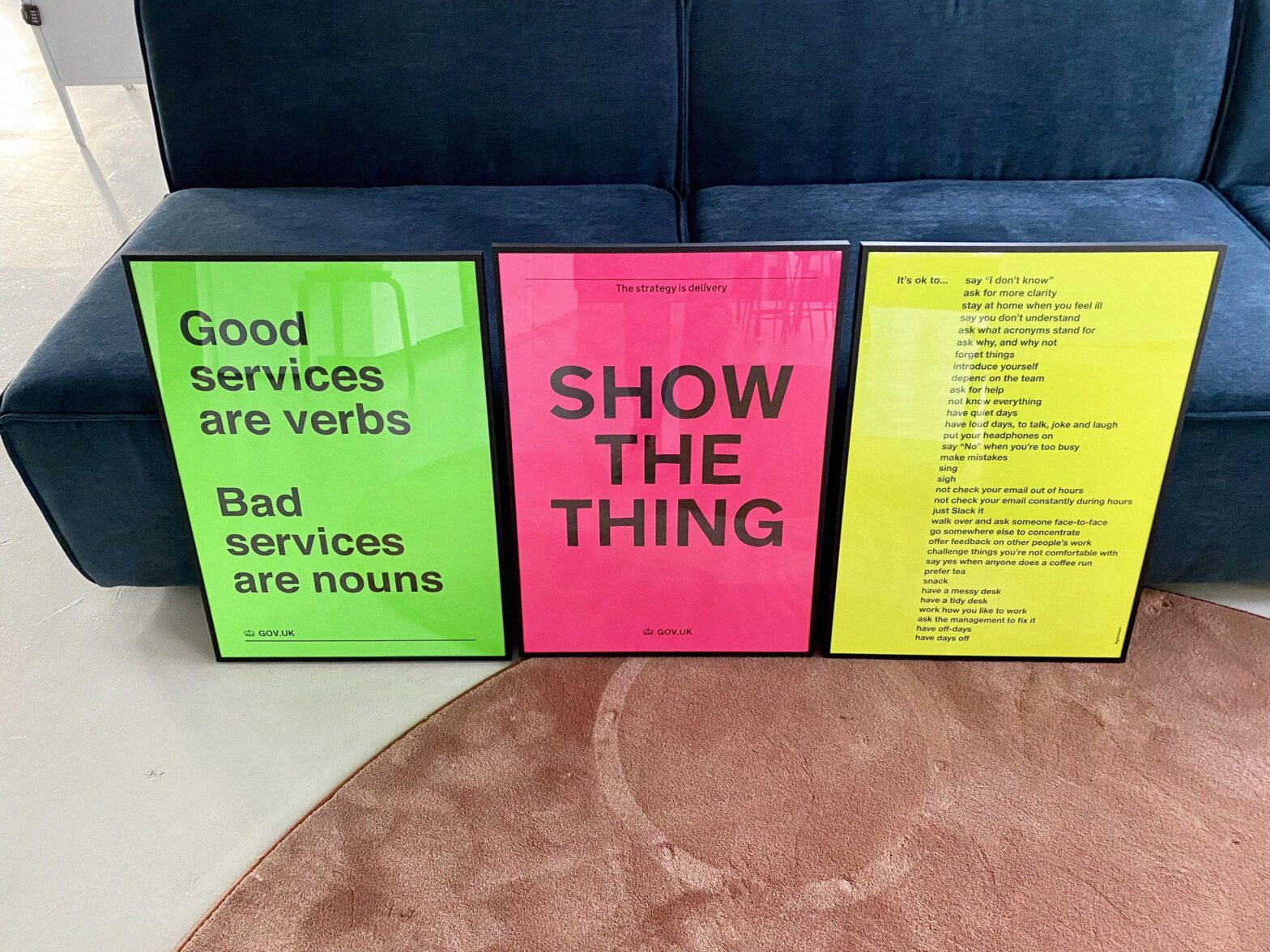 3 framed posters standing in front of a big sofa. 1st poster says: Good services are verbs, bad services are nouns. 2nd poster says: Show the thing. 3rd poster says: It’s ok to – and then lists a long list of things that are ok in the work culture, including OK to say I don’t know, ask for more clarity, ask for help