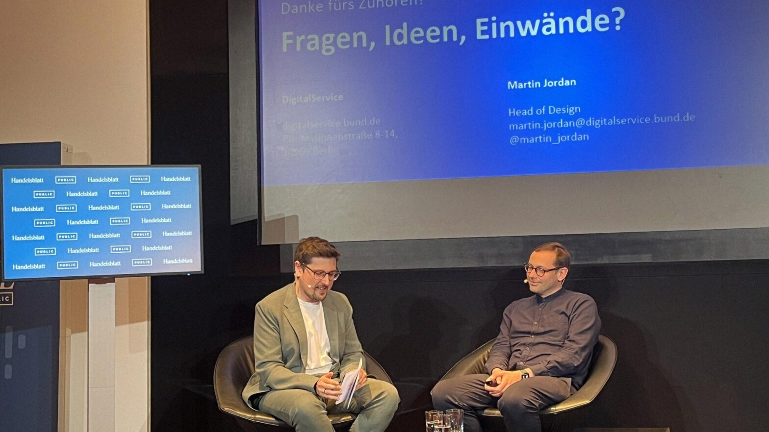 Two young-ish white men with glasses on stage talking to each other with a presentation slide above them saying ‘Questions, ideas, concerns?’ in German