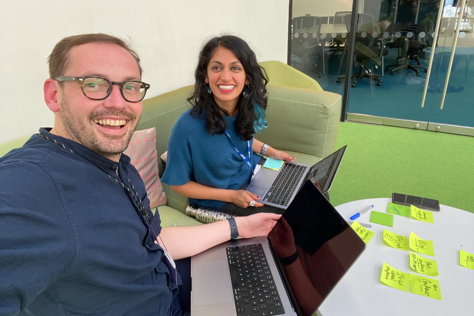 Selfie of Martin Jordan and senior user researcher Paloma Jain in a modern office space sitting on a comfy sofa with laptop computers and stickie notes
