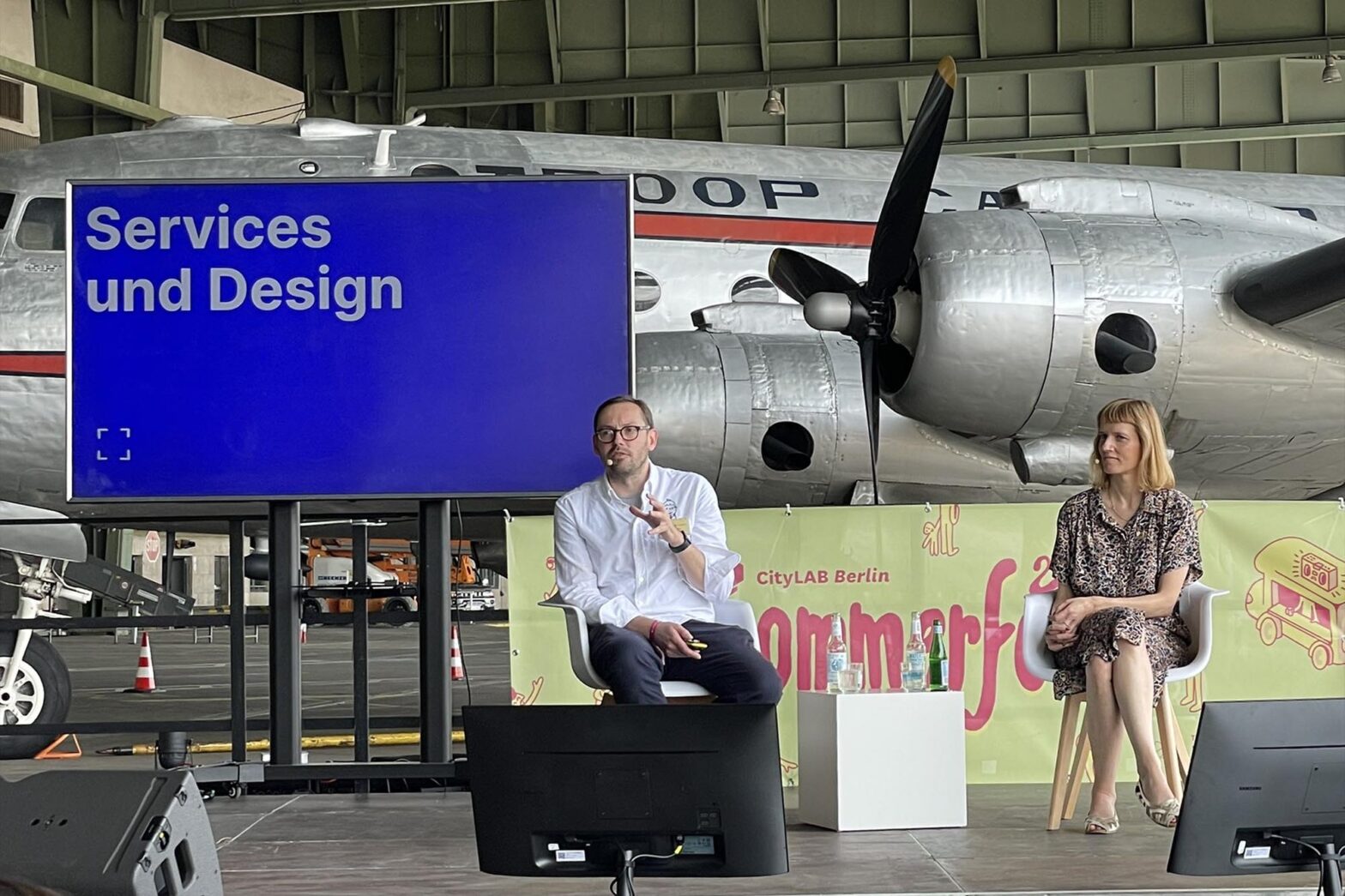 Two white people, a man and a woman, on a conference stage with a big screen behind them saying ‘Services and Design’ in German; an airplane is standing right behind them, a banner in the background says ‘CityLAB Berlin summer festival’