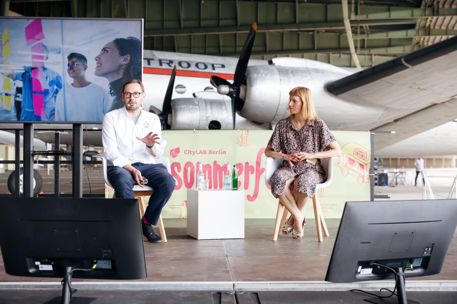 A man and a woman on a stage talking with an old airplane standing behind them