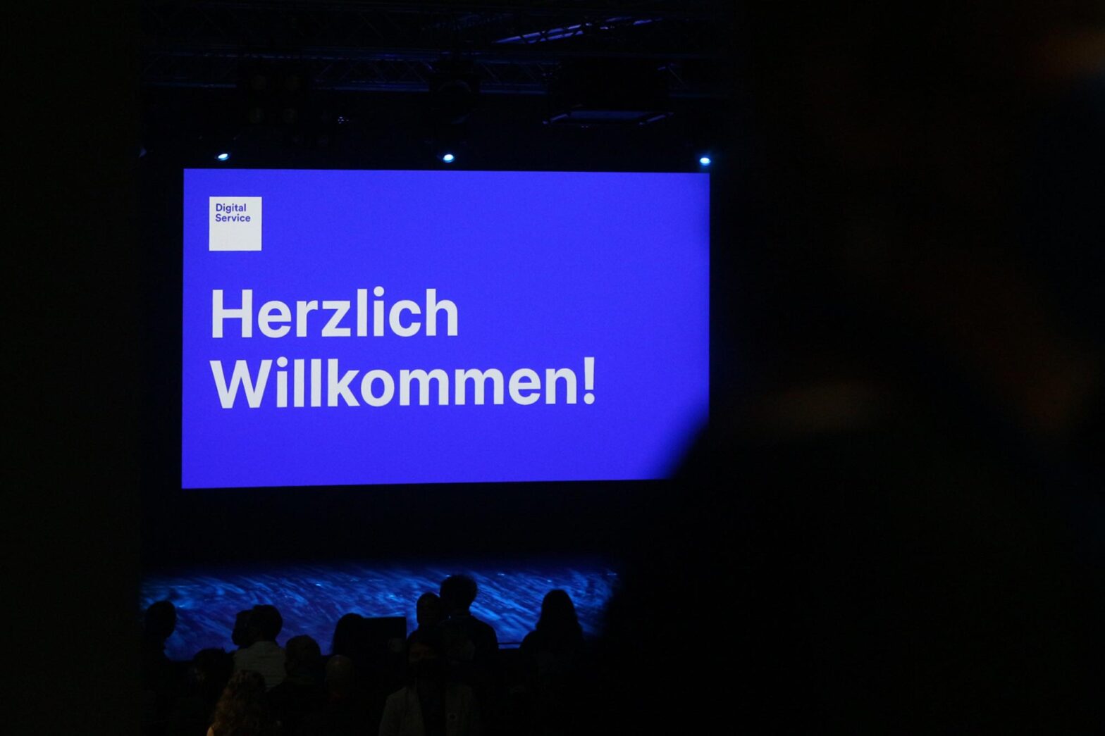 A stage with a huge projected slide saying ‘A warm welcome’ in German – with a logo of DigitalService