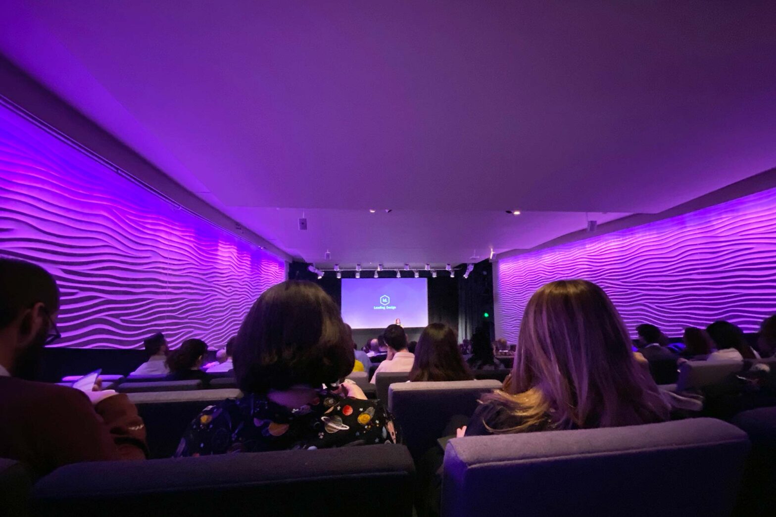 A cinema-like room with purple light, people sitting in many rows, 2 people with longer hair just in front of the camera and a small logo is projected at the end of room saying LD – leading design