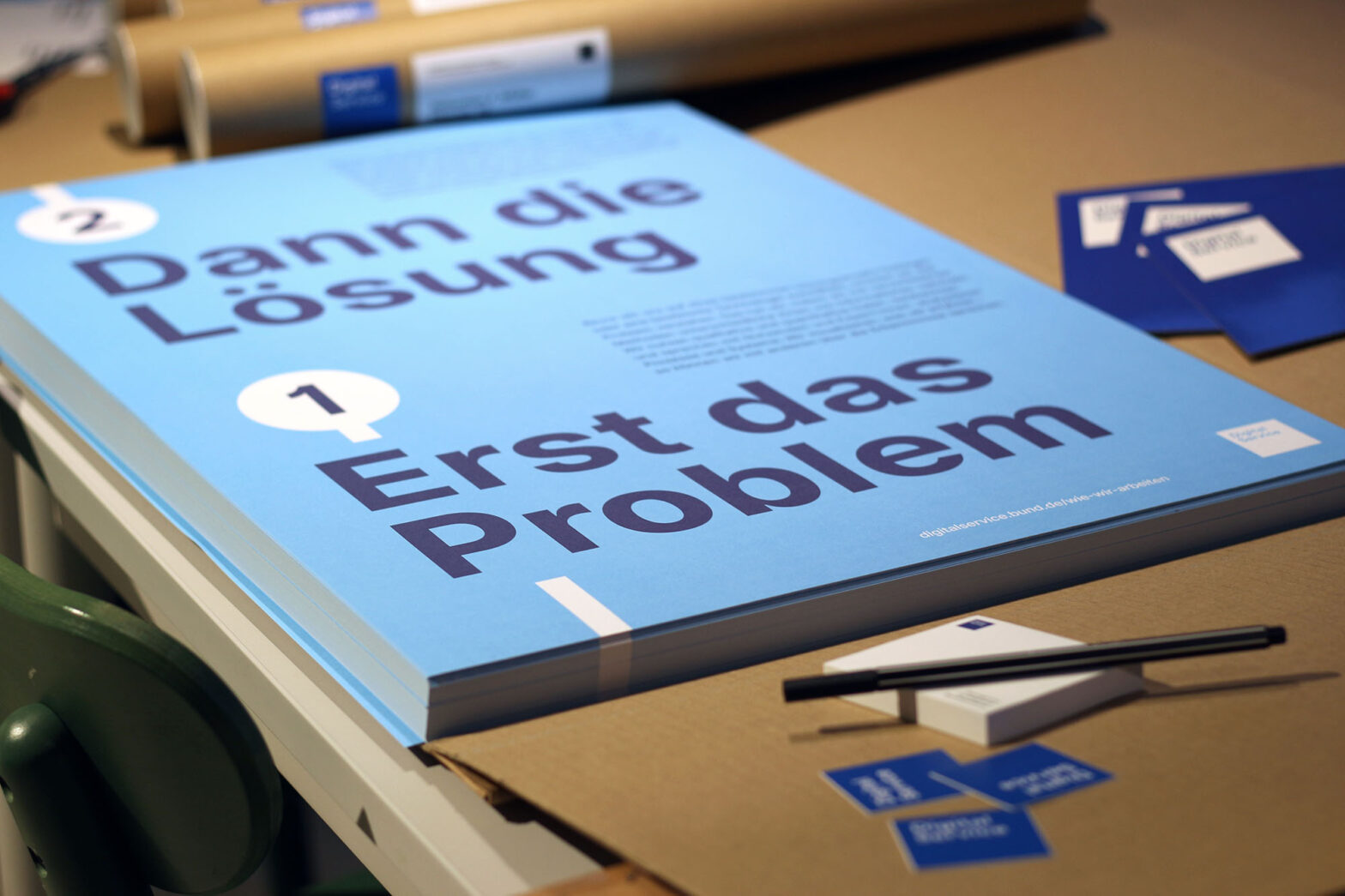 A pile of light blue posters surrounded by mailing tubes, stickers, post-it pads and blue cards all carrying the square logo of the Digital Service; the poster says in German: “Solution second, problem first”