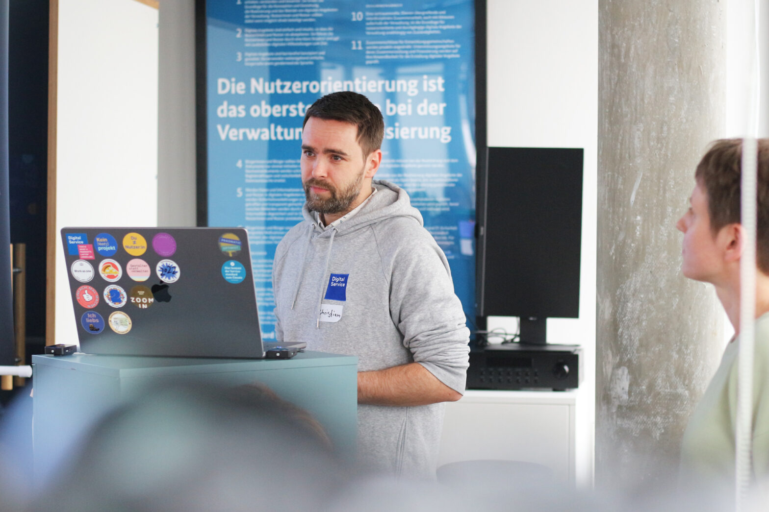 A youngish white man in a grey branded hoodie saying “Digital Service” standing in front of a podium with a computer with lots of colourful stickers; in the background is a poster that reads “User-centricity is the key principle when digitalising administration” – a woman with shorter hair is standing on the side looking at him