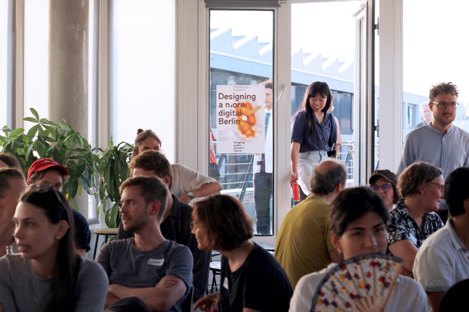 People sitting in an open office space with plants – in the background a younger woman of Asian descent entering from a terrace door with a poster hanging next to it: it shows a pixelated curry sausage and says ‘Designing a more digital Berlin’