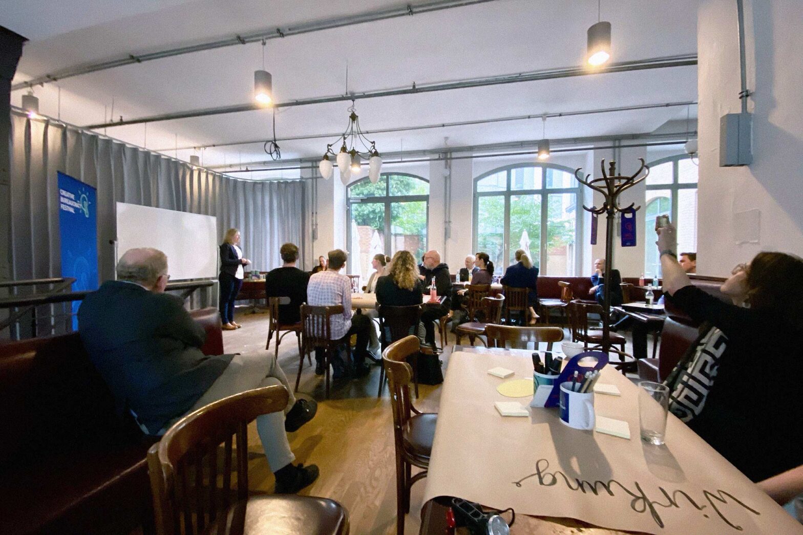 A group of people in a wide and bright cafe-like place with various tables and classic coffee house chairs – one person is standing in front of a whiteboard