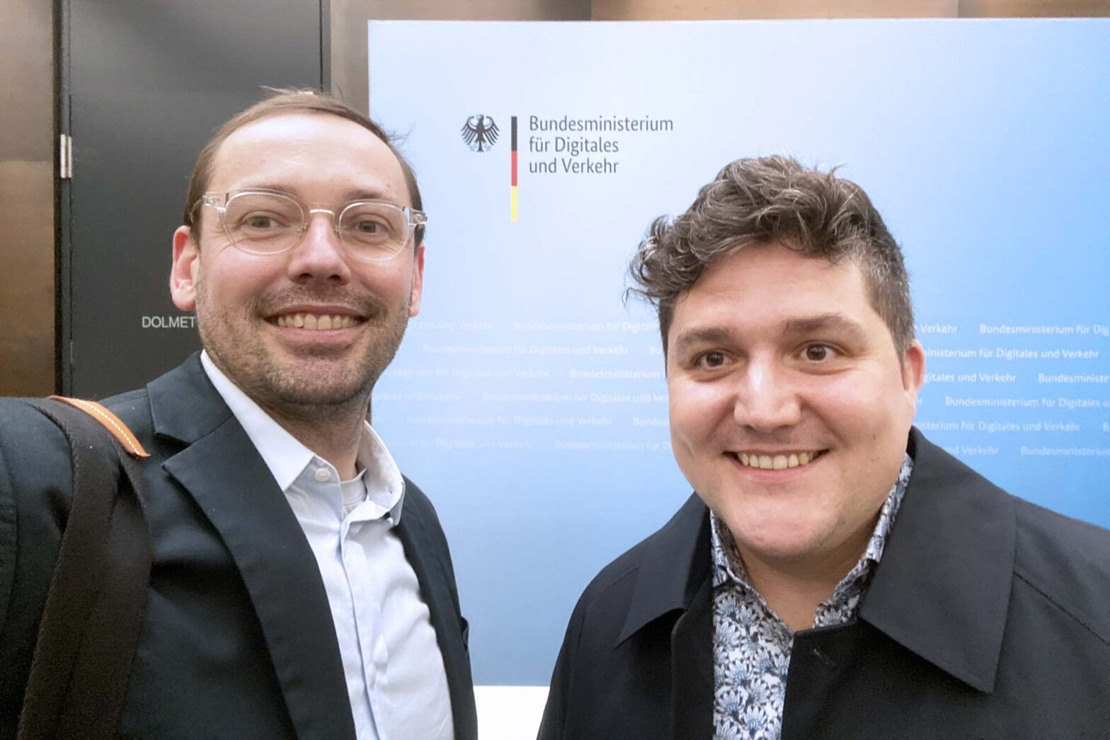 Two middle-aged white men in casual attire in front of a wall with the logo of the German federal ministry of digital and transport – both are smiling into the camera