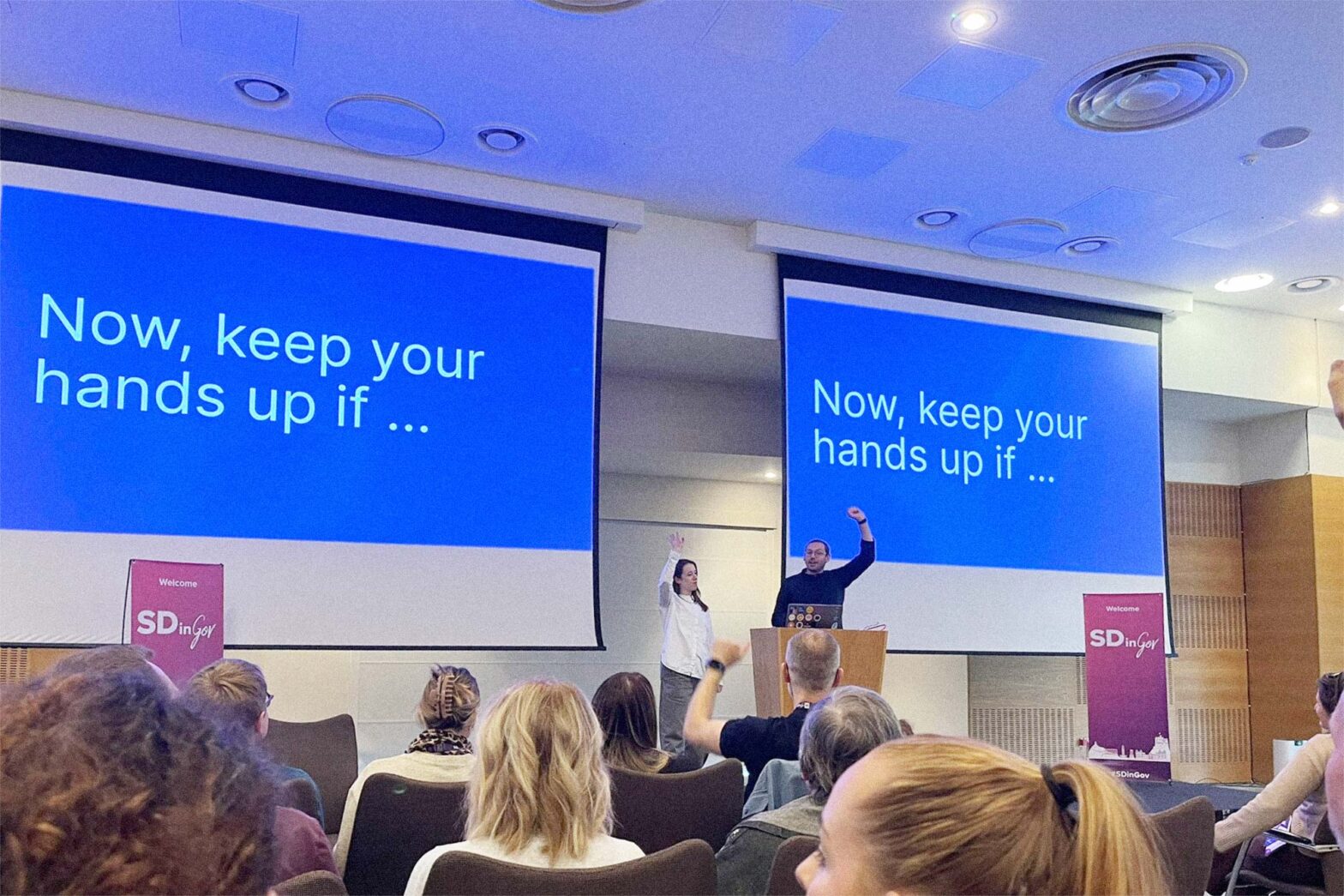 2 people, a woman and a man, speaking in a bigger space with wooden panels in front of a dozen people – both speaker raise their hands; slides in the background say" “Now, keep your hands up if …”