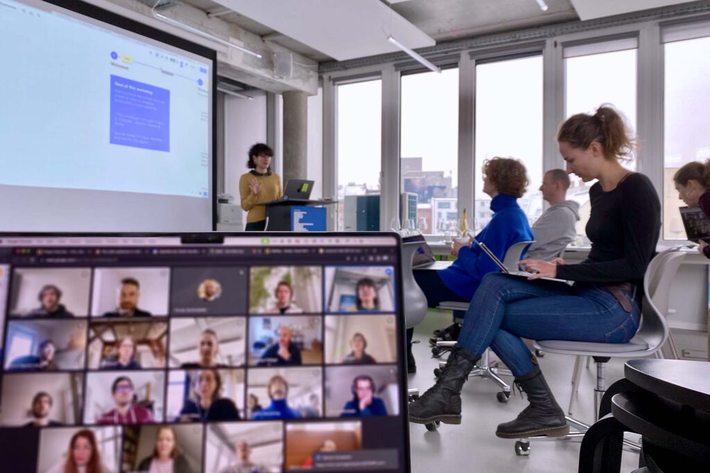 5 people in a well-lit open office space with 1 person standing next to a big projected screen next to a lectern – and an out of focus laptop screen with 19 more people in the foreground