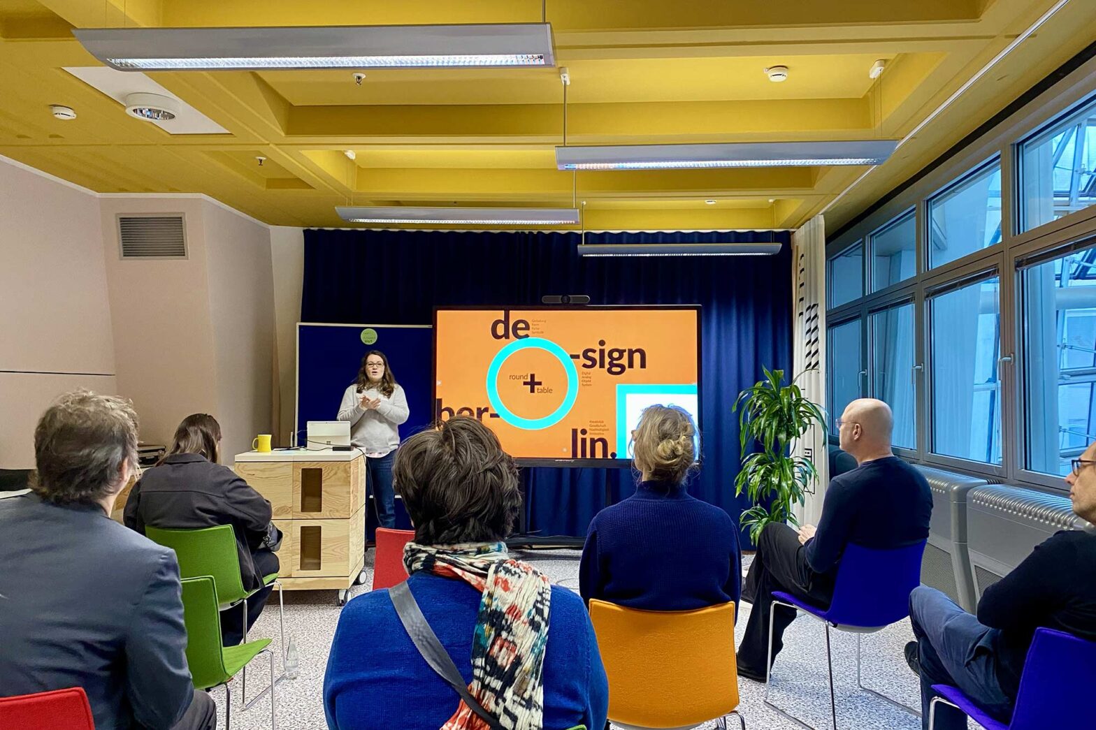 A woman with light skin and long hair in a modern colourful space speaking in front of 6 sitting people – next to her is a big TV set saying Berlin Design