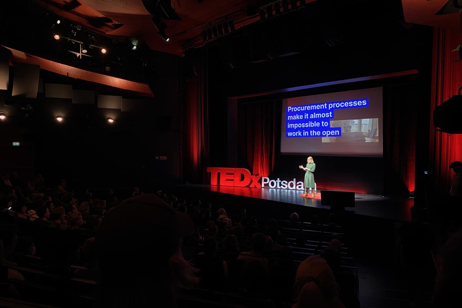 A white woman with ginger hair in a minimal green dress and brown leather boots standing on a big all-black stage on a red circular rug – next to her are the letters TEDx standing as red letters, behind her is a projected slide