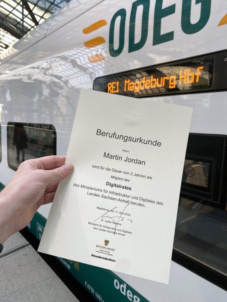 A light-skinned hand holding a certificate of appointment for Martin Jordan for the Digital Council of the Ministry for Infrastruktur and Digital in the Federal State Sachsen-Anhalt with signature of the minister in front of a train train displaying its destination Magdeburg