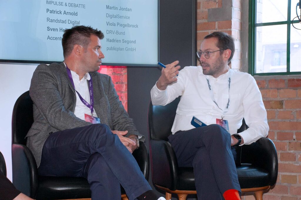 Two men are seated in black chairs, engaged in discussion. The man on the left wears a gray blazer and a purple lanyard. The man on the right, wearing glasses and a white shirt, is gesturing with a pen. Behind them, a screen displays names and affiliations, and the background features a brick wall and a window.