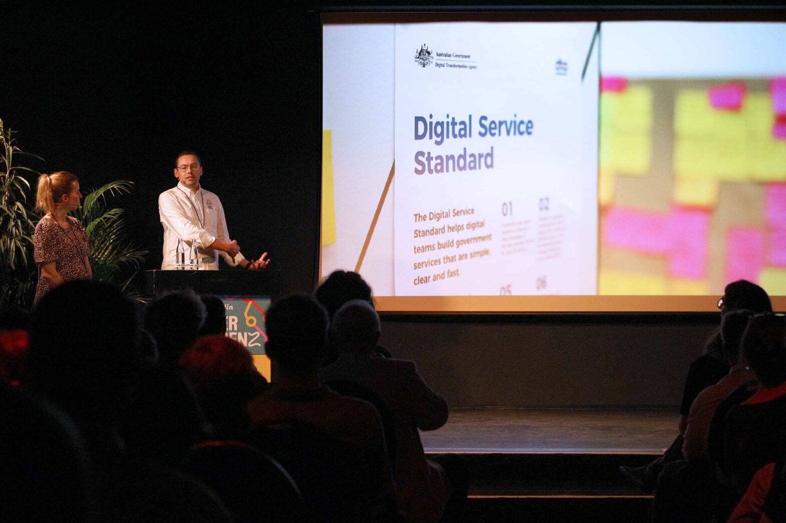 Two presenters stand on a stage in a dimly lit auditorium, facing an audience. The man, wearing glasses and a white shirt, gestures while speaking. The woman, dressed in a patterned dress, stands beside him. Behind them is a large screen displaying a slide titled "Digital Service Standard" with text explaining its purpose to help digital teams build government services.
