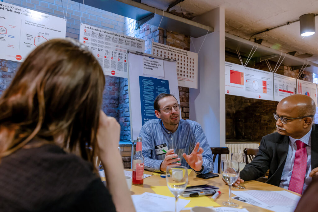 Three people are seated at a table in a meeting room, engaged in discussion. The man in the center, wearing glasses and a striped shirt, gestures with his hands while speaking. A man to his right, who has dark skin, wears glasses and a suit with a pink tie and listens intently. A woman with brown hair, seen from the back, is on the left side of the image. The table has glasses, papers, and a lemonade bottle. Behind them, various informational posters and charts are displayed on the walls, which are made of exposed brick.