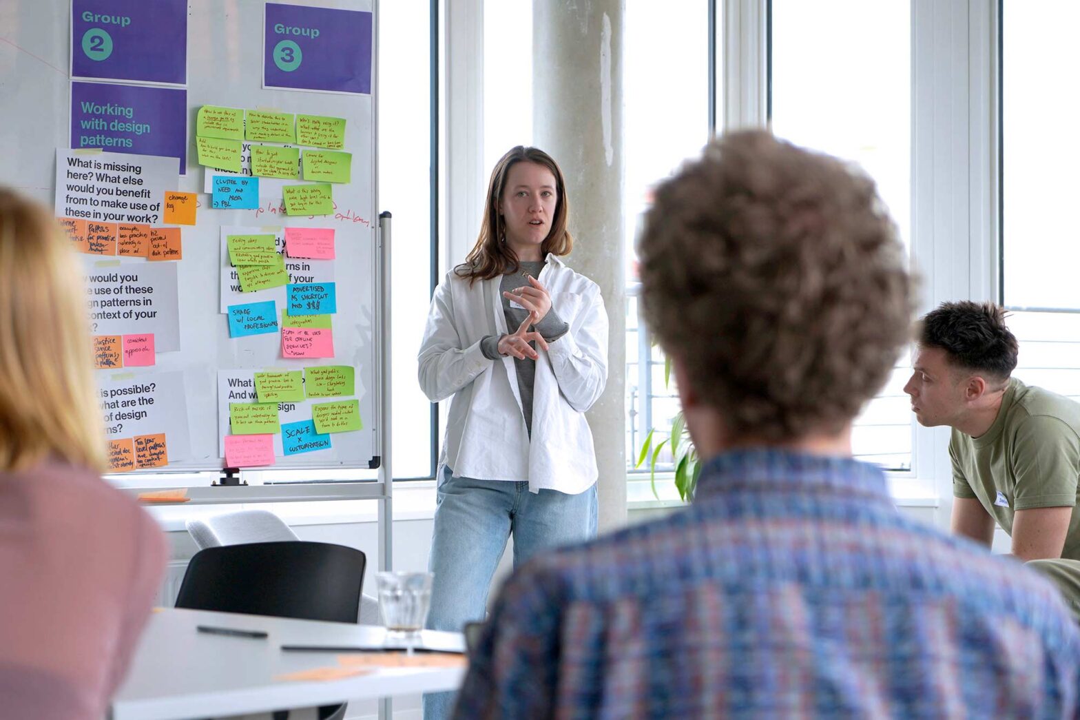A woman stands in front of a whiteboard filled with colorful sticky notes, presenting to a group. She wears a white shirt over a gray top and blue jeans, gesturing with her hands. Two individuals, one with curly hair and another in a green t-shirt, listen attentively. The whiteboard has two sections labeled "Group 2" and "Group 3" with various questions and comments written on the notes. The room is bright with large windows in the background.