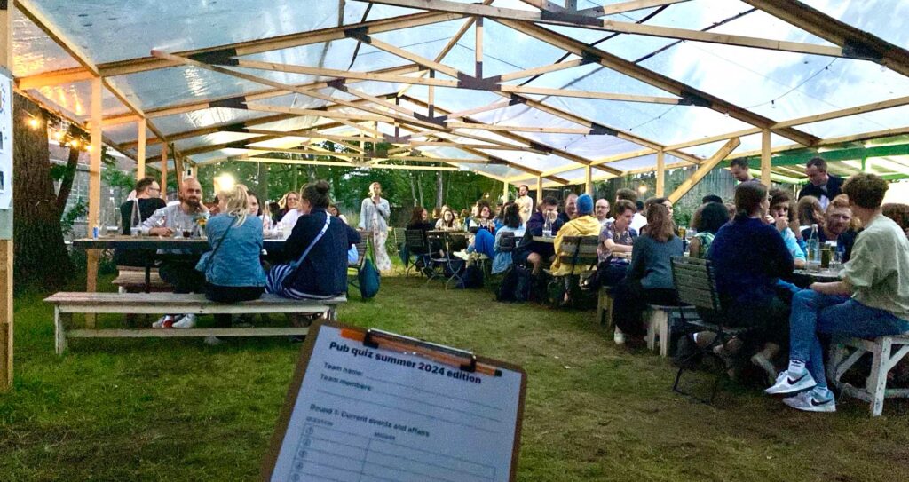 A large group of people sits under a transparent tent with wooden supports, at picnic tables and benches in an outdoor setting with trees. The scene is well-lit with string lights. In the foreground, a clipboard with a "Pub quiz summer 2024 edition" sheet is visible, indicating a quiz night. People are engaged in conversations, eating, and drinking, creating a lively atmosphere.