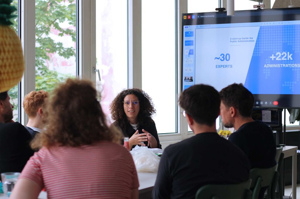 A woman with curly hair and glasses is leading a meeting, speaking to a group of five people seated around a table. She gestures with her hands as she talks. Behind her, large windows offer a view of greenery outside. A large screen to her right displays a presentation slide with text reading "~30 EXPERTS" and "+22k ADMINISTRATIONS." The attendees, whose backs are facing the camera, include individuals with different hair colors and styles. The room is bright, with natural light streaming in from the windows.