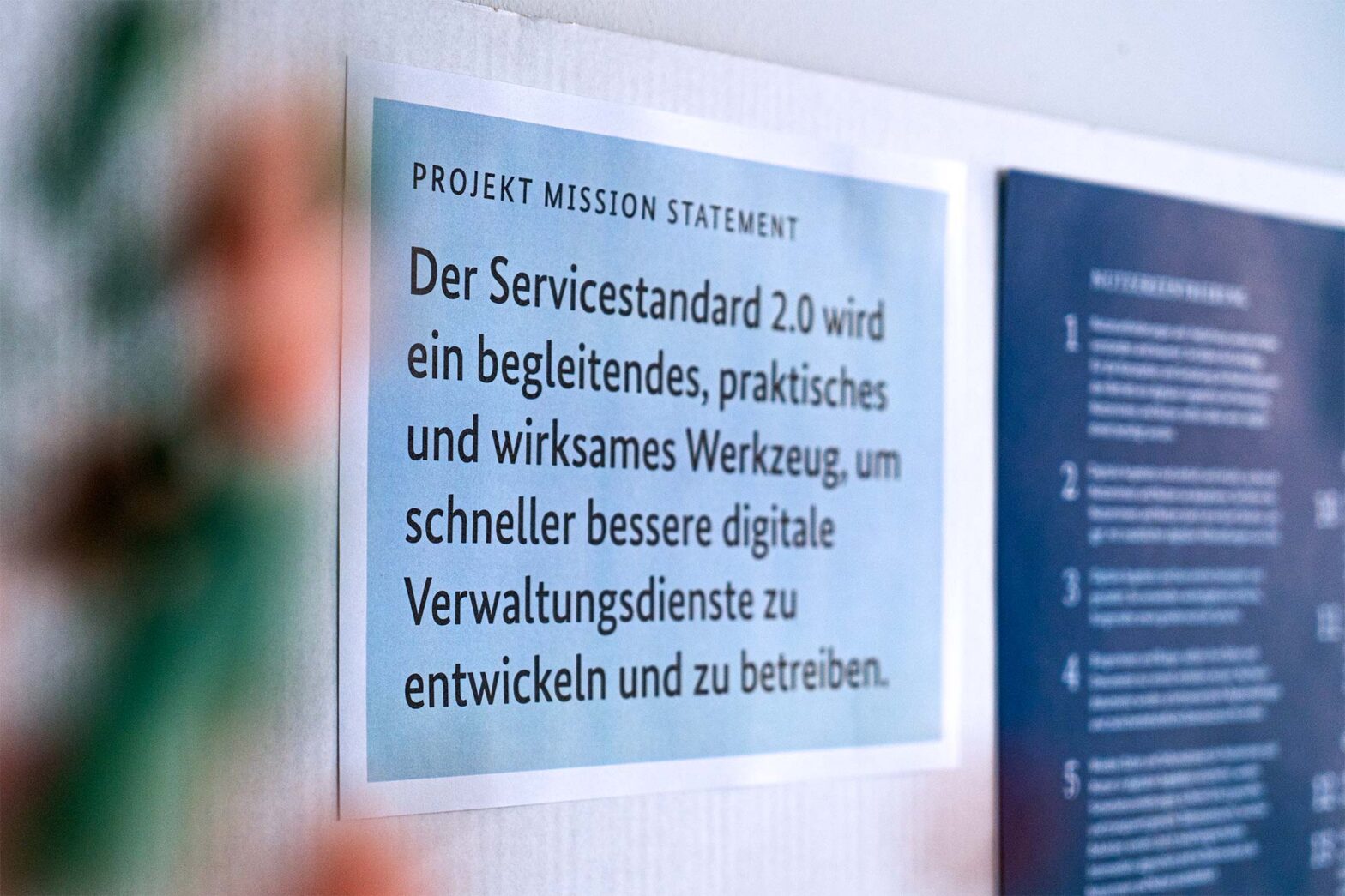 A sheet on a card board next to a poster with a numbered list of text blocks; the sheet says in German: “The Service Standard 2.0 will be an enabling, practical and impactful tool to develop and operate better digital public services faster.”