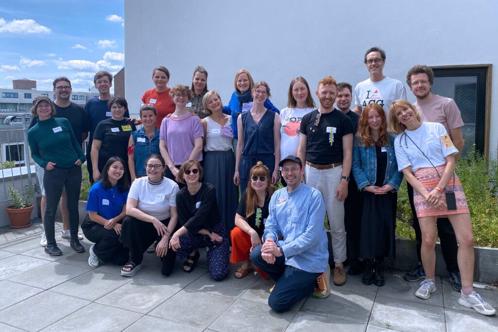 A group of twenty-one people poses for a photo on a rooftop terrace with a clear blue sky in the background. They are casually dressed and wear name tags. Some are standing, while others are kneeling or sitting in front. They smile at the camera, creating a friendly and relaxed atmosphere. The terrace has a potted plant and railing, with buildings visible in the distance.