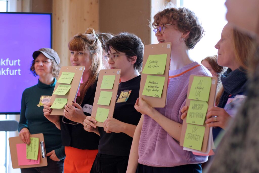 Five women stand indoors, each holding a clipboard with green sticky notes listing various topics. They are smiling and appear engaged in a workshop or presentation. One woman wears a green top and cap, while the others are dressed casually in colorful clothing. A purple screen in the background displays partial text reading "Frankfurt." The atmosphere is collaborative and focused.