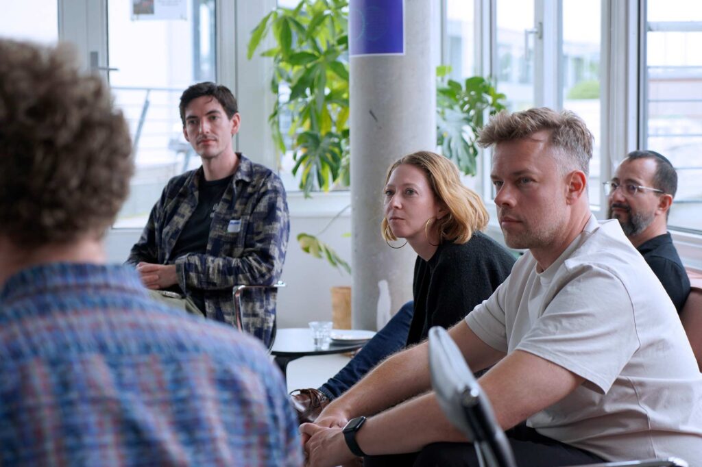 Four people sit attentively in a meeting room, listening to someone outside the frame. The man on the left wears a plaid shirt, the woman next to him has short blonde hair and large hoop earrings, the man beside her is in a light-colored shirt, and the man in the back wears glasses and a dark shirt. There are large windows and a potted plant in the background, providing a bright, natural light to the room. A man with curly hair is partially visible in the foreground.
