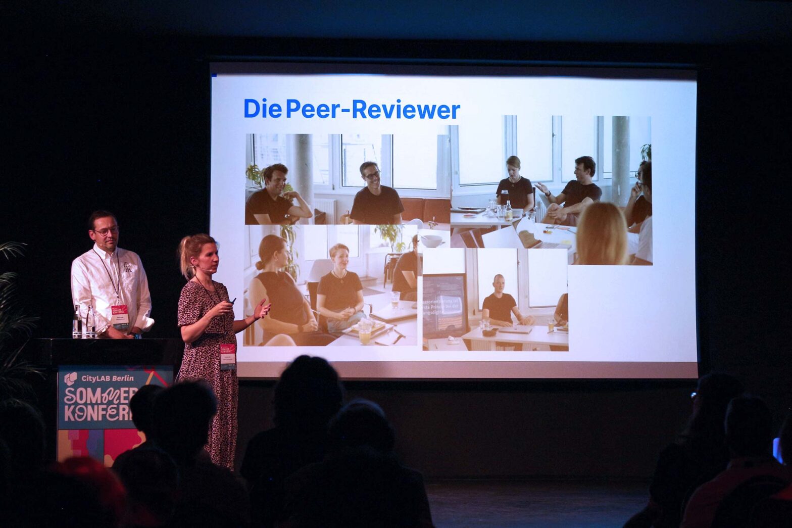 A man and a woman are presenting on stage in front of an audience. Behind them, a large screen displays a slide titled ‘The peer reviewers’ in German with four photos showing people sitting around a table engaged in discussion