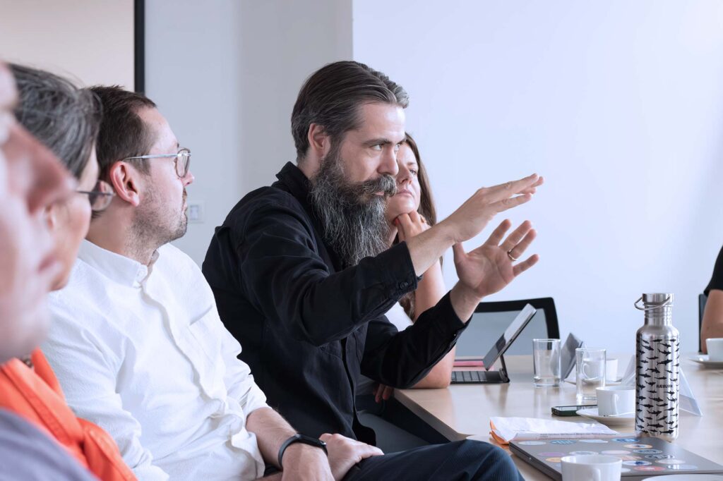 A group of people sit around a conference table during a meeting. A man with a long beard, wearing a black shirt, is speaking and gesturing with his hands, while others listen attentively. A metal water bottle and some notebooks are on the table.
