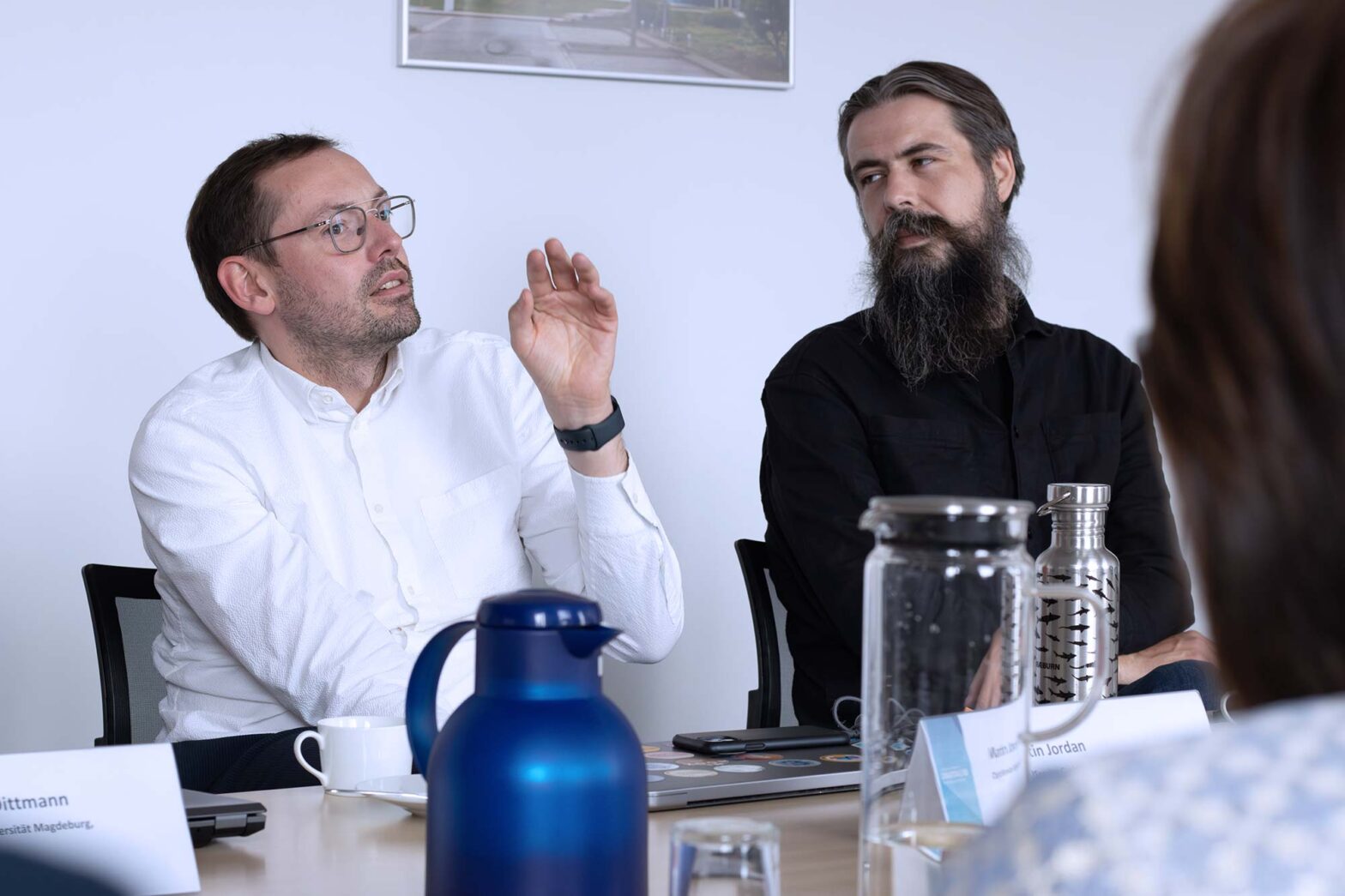 Two light-skinned men are seated at a meeting table, engaged in conversation. The man on the left, wearing glasses and a white shirt, gestures with his hand while speaking. The man on the right, with a long beard and dark shirt, listens attentively. In the foreground, out-of-focus objects, including a blue thermos and name cards, are visible.