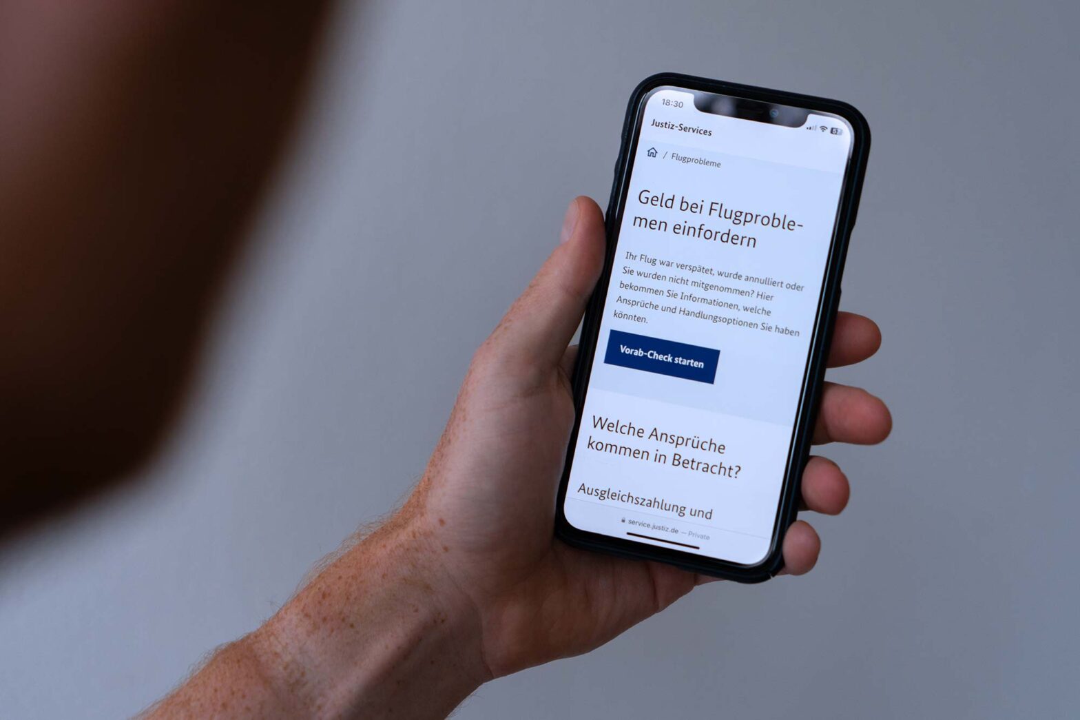 A hand holding a smartphone displaying a German online form related to flight issues. The screen reads, "Claim money for flight problems." Below this, it explains that if the flight was delayed, canceled, or overbooked, users can find information on their rights and options. There is a blue button labeled "Start preliminary check".