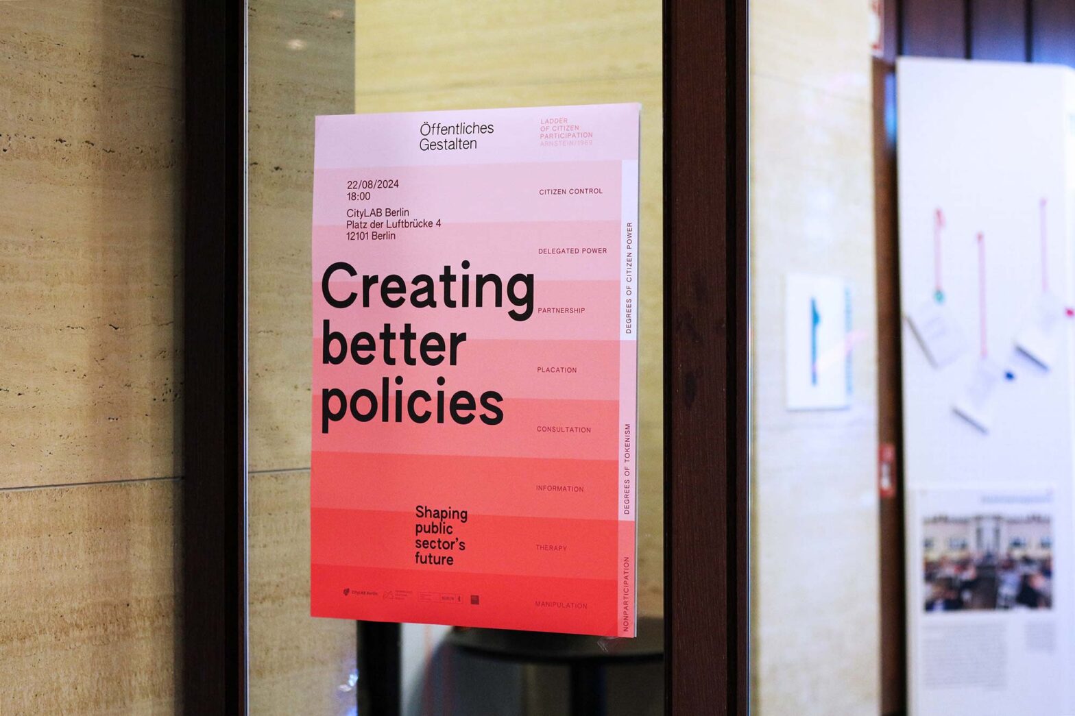 A poster on a wall promoting an event titled "Creating Better Policies." The poster has a gradient from pink to red and lists information such as the date, time, and location: 22/08/2024 at CityLAB Berlin. The right side of the poster displays a "Ladder of Citizen Participation," with levels ranging from "Citizen Control" to "Manipulation."