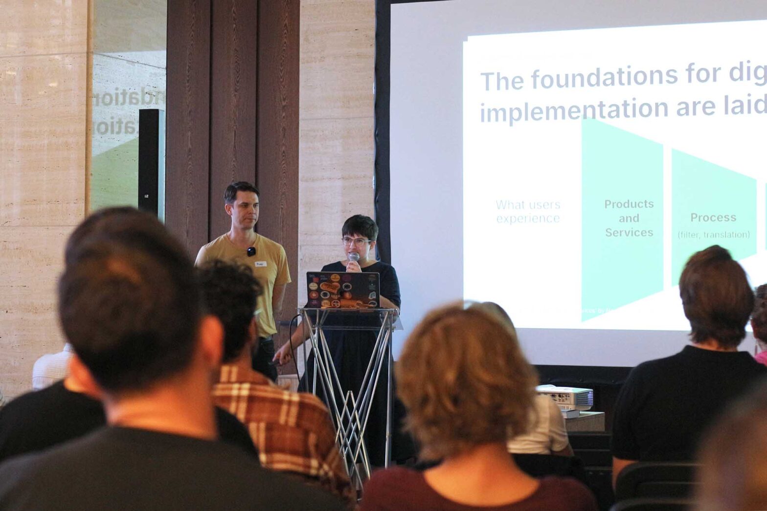 Two presenters stand at the front of a room facing an audience, one holding a microphone and the other standing beside them. A presentation slide is projected on a screen behind them, displaying the title "The foundations for digital implementation are laid" with sections labeled "What users experience," "Products and Services," and "Process." The audience is seated, with heads and shoulders visible in the foreground.