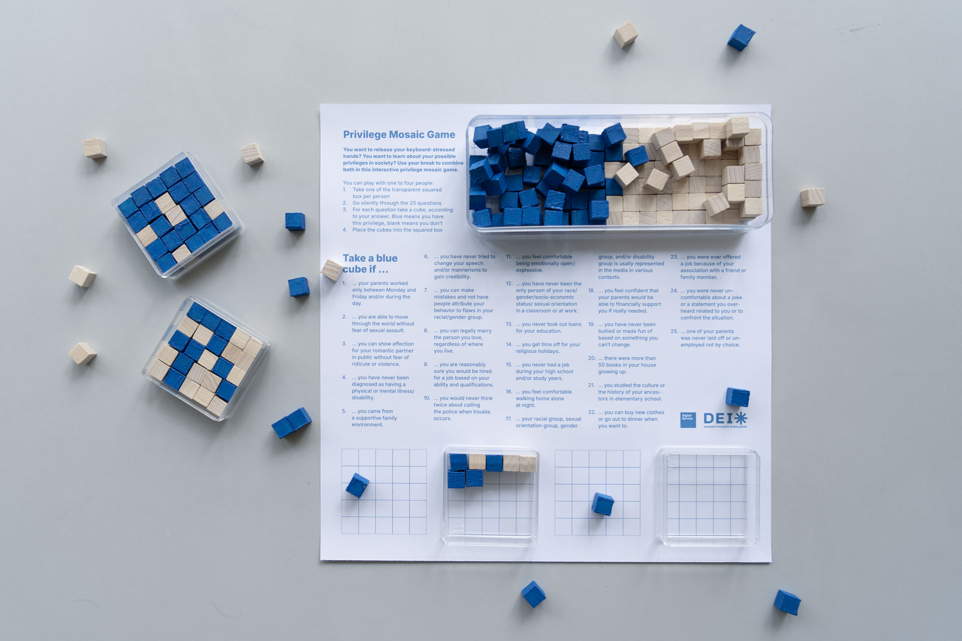 An educational game setup titled "Privilege Mosaic Game," featuring blue and beige wooden cubes. The cubes are arranged in plastic trays with grid patterns. An instruction sheet lists scenarios that indicate privilege, prompting players to take a blue cube if applicable. The game elements are scattered on a white surface.